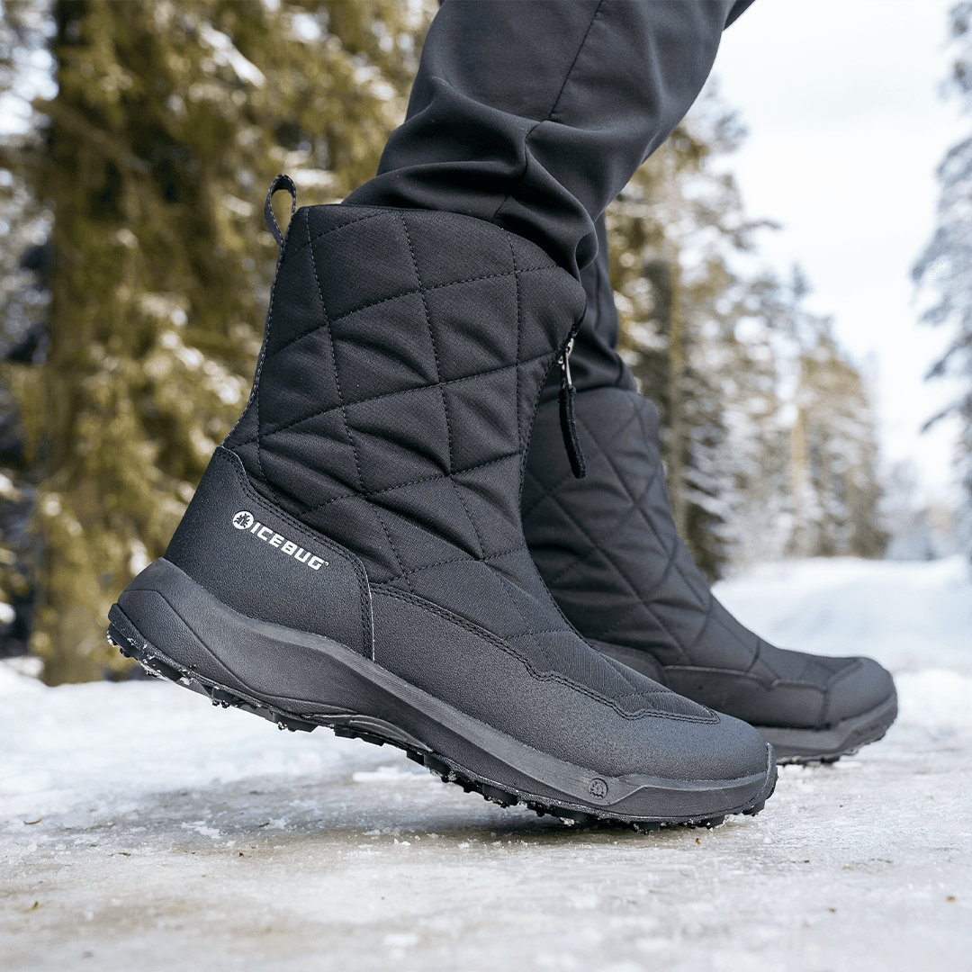 Close-up of black quilted winter boots on a snowy path, surrounded by tall evergreen trees.