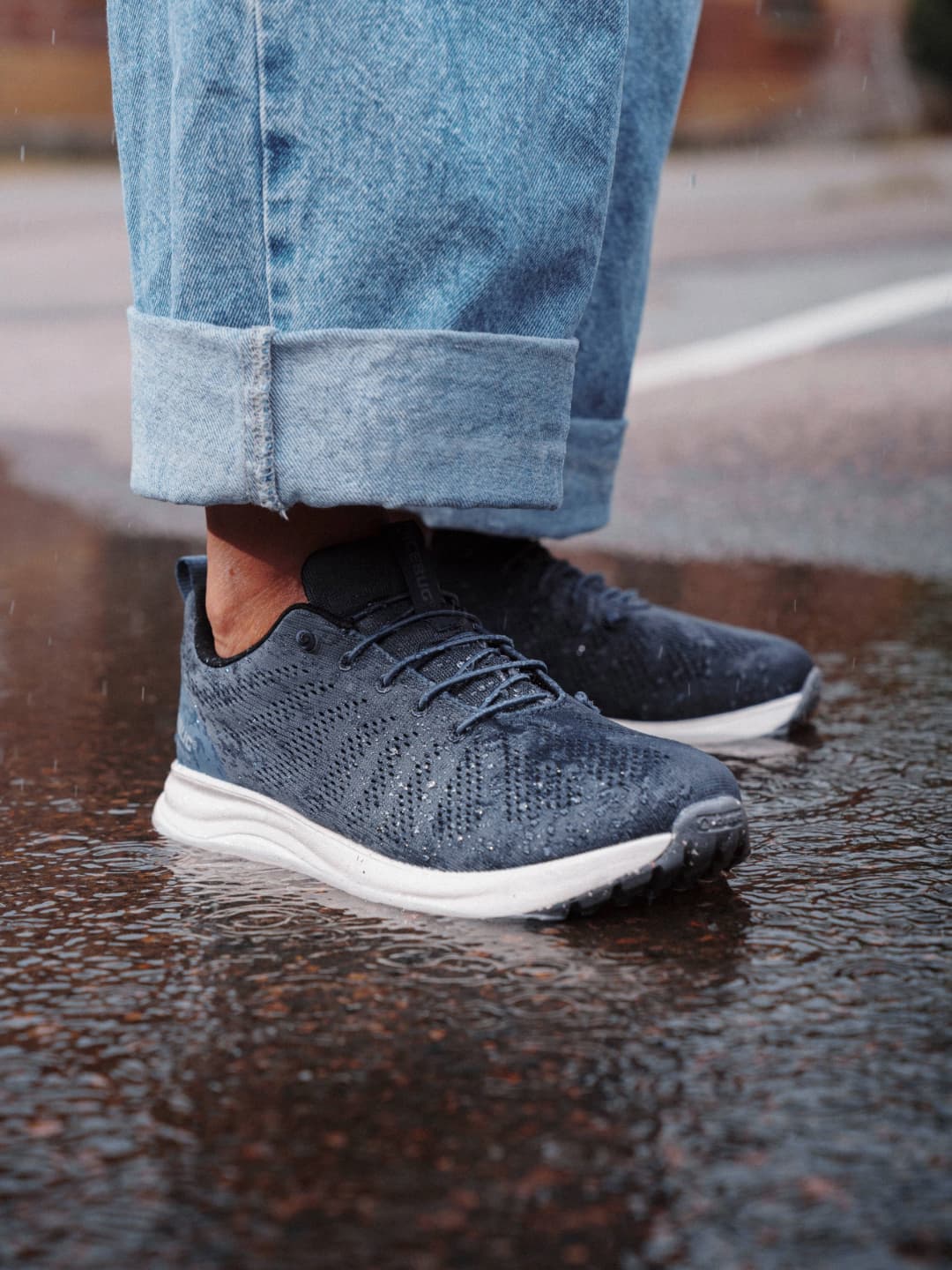 Person wearing blue jeans and dark blue sneakers standing on wet pavement, reflecting the shoes and cuffs of the jeans.