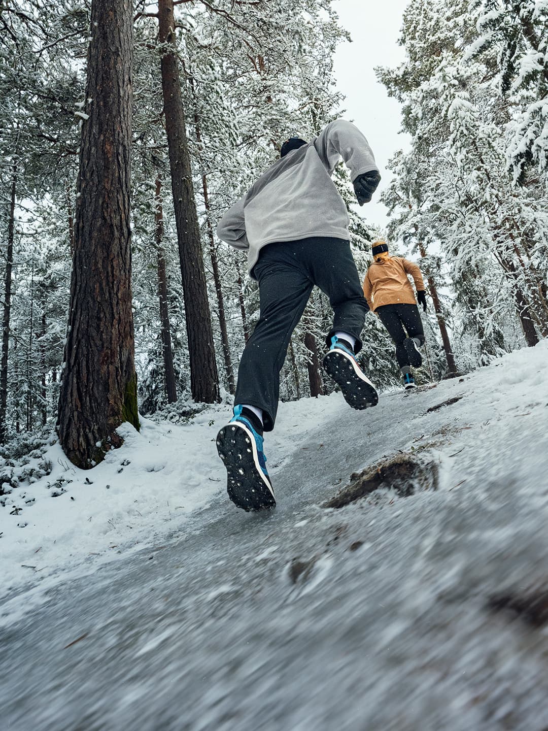 Two people running uphill on a snowy forest trail, wearing winter gear and Icebug studded winter running shoes. Snow-covered trees surround them.