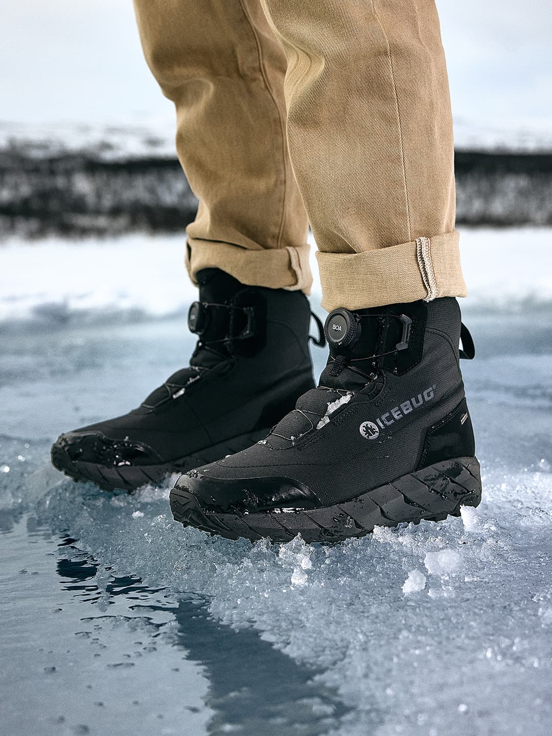 Person wearing black Icebug winter boots and beige pants standing on icy surface.
