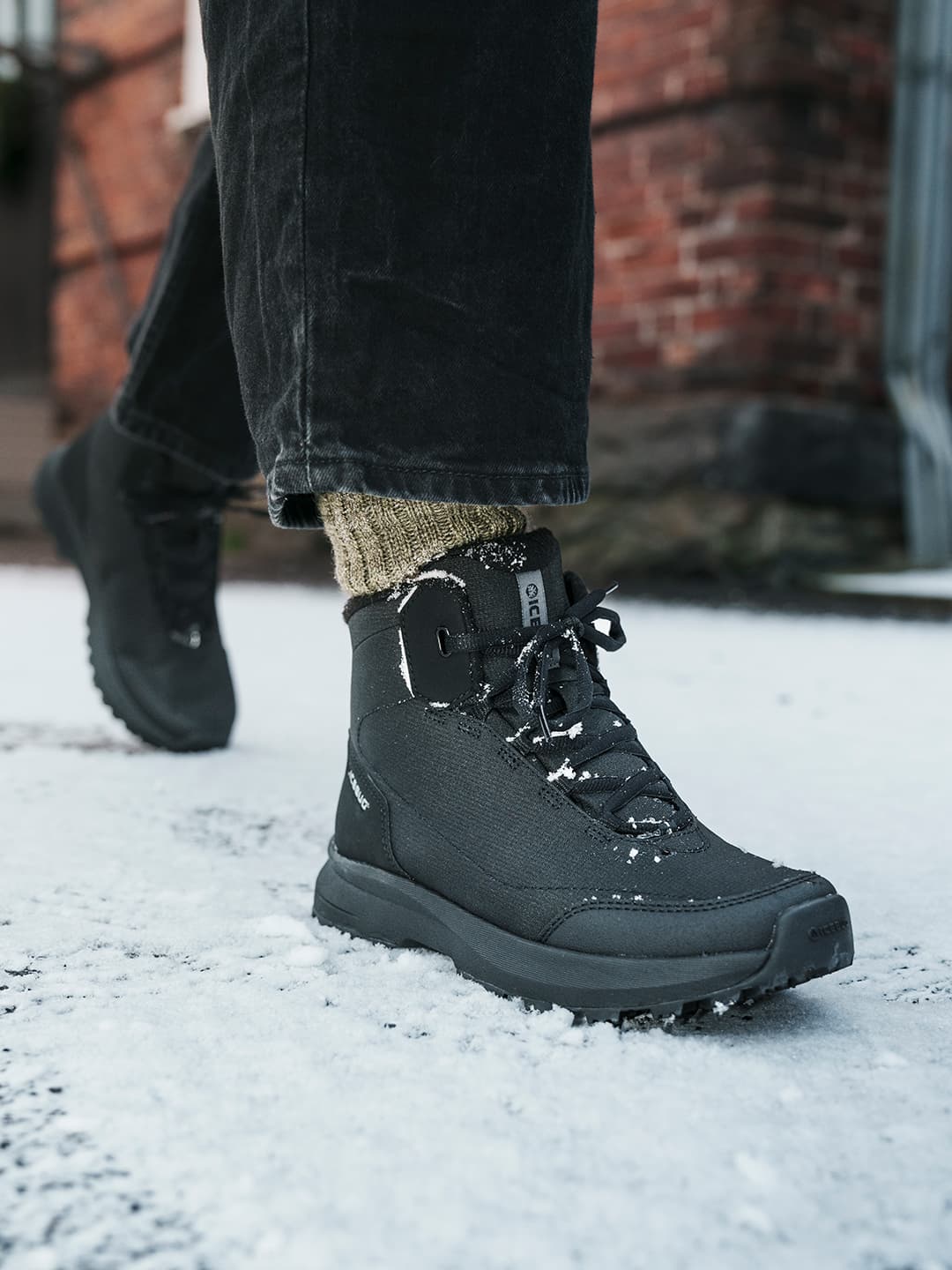 Person wearing black boots and dark pants walking on snow-covered ground, with a brick building in the background.