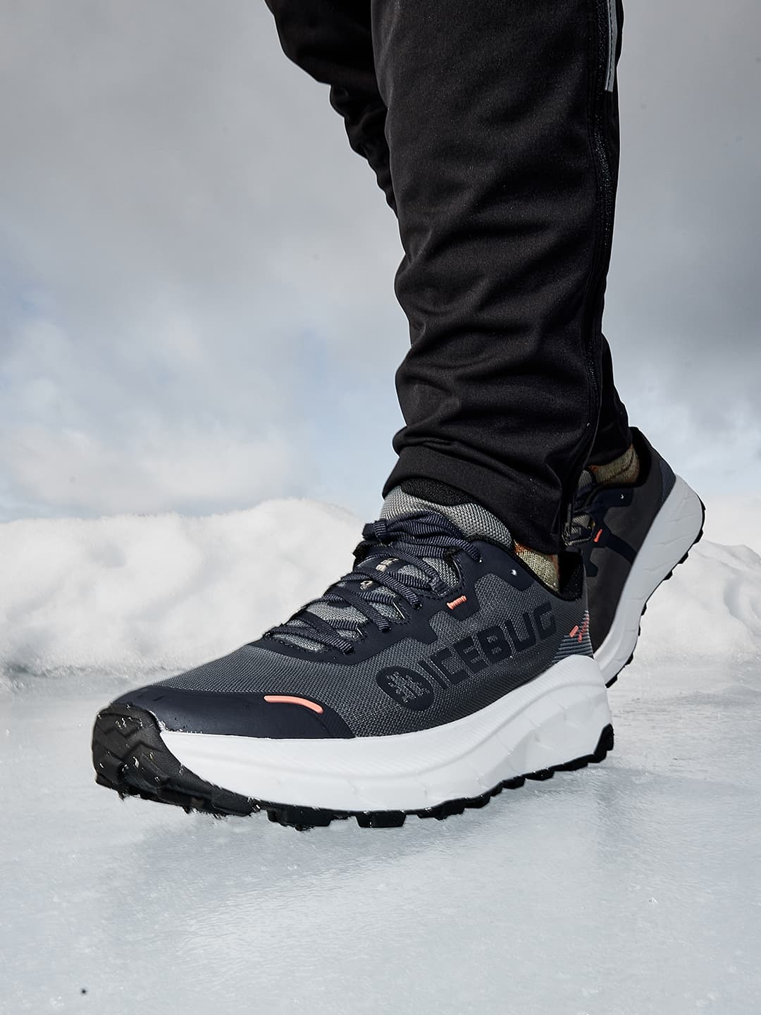Close-up of a person wearing grey Icebug winter running shoes with white soles, standing on icy ground against a cloudy sky backdrop.