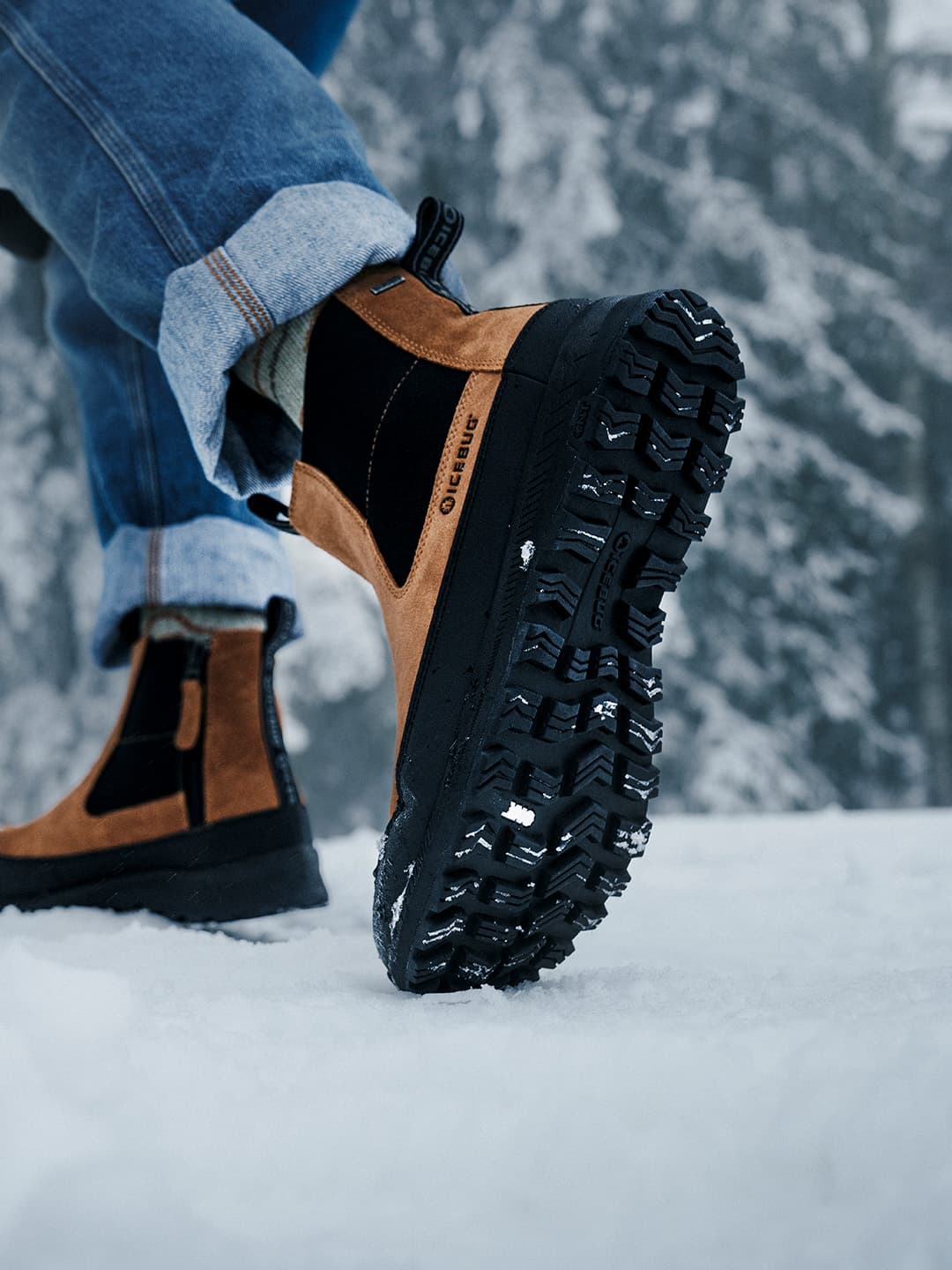 Close-up of a person wearing brown and black winter boots with thick treads, walking on snow. Denim jeans are slightly rolled up.