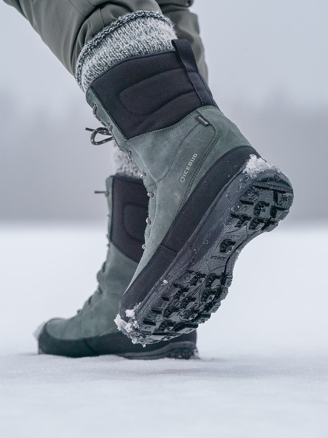 Close-up of a person walking on snow, wearing green and black winter boots with visible tread and snowflakes on the soles.
