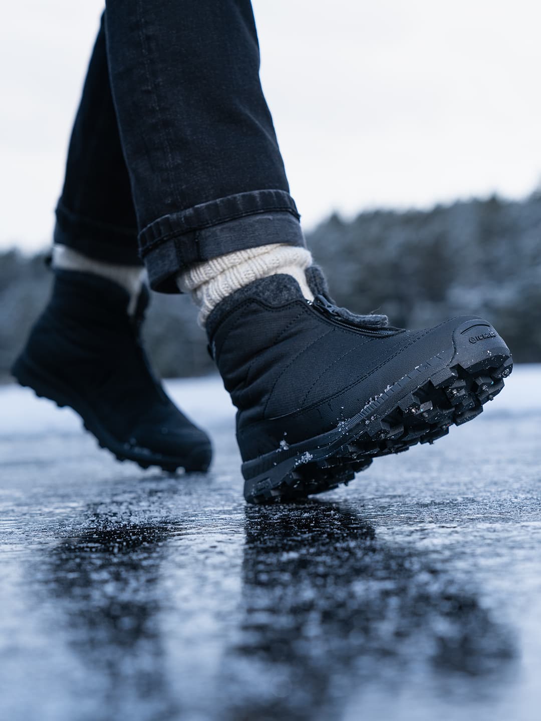 Close-up of a person walking on an icy surface, wearing black winter boots, dark jeans, and cream-colored socks.