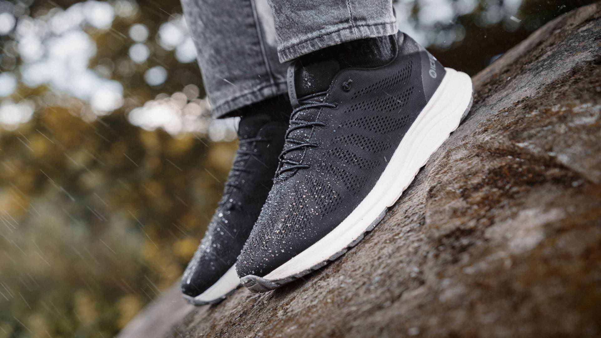Person wearing black sneakers with white soles standing on a wet rock, surrounded by a blurred natural background.