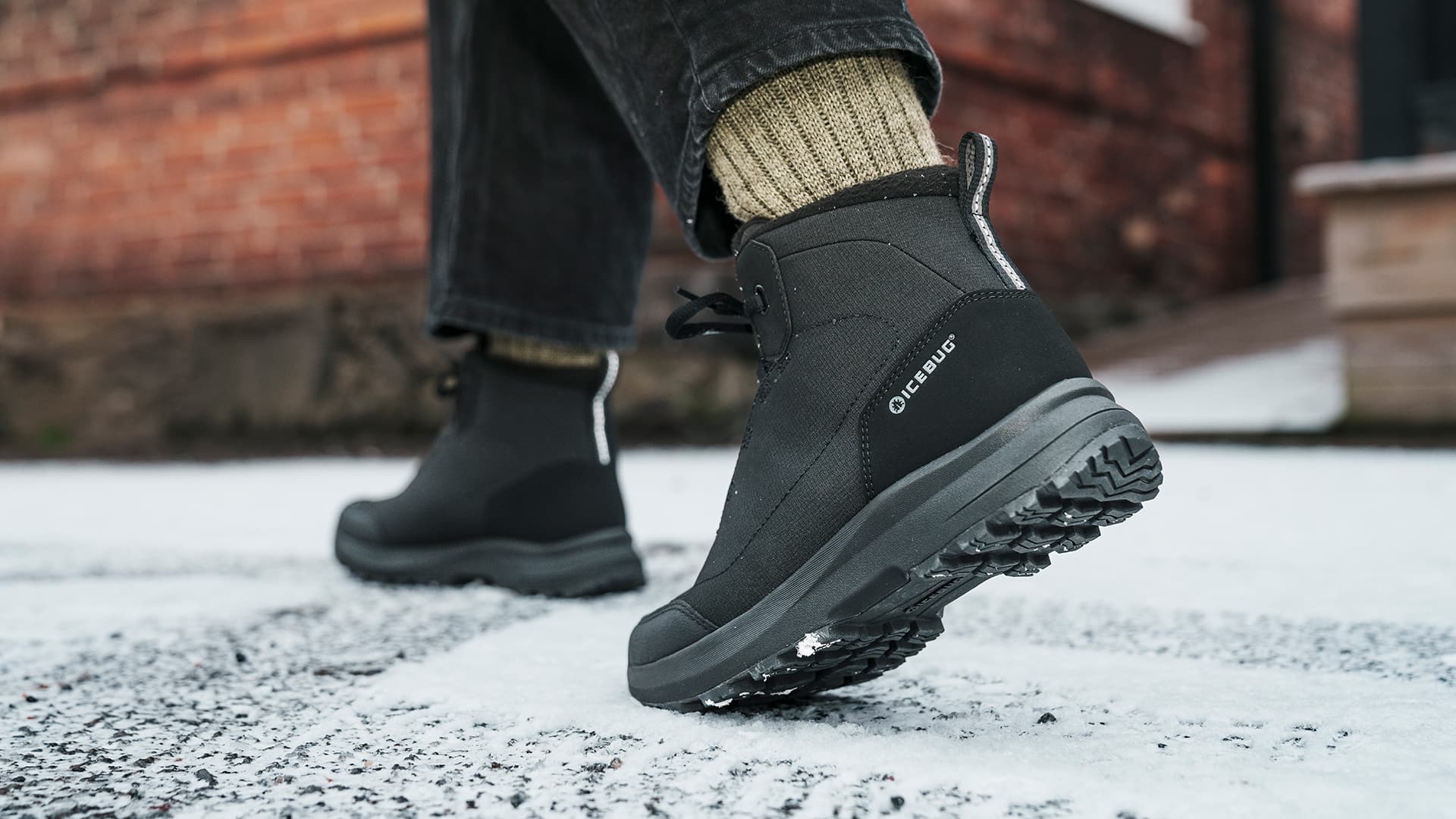 Close-up of a person wearing black winter boots walking on a snowy, icy pavement. The boots have a rugged sole for traction.