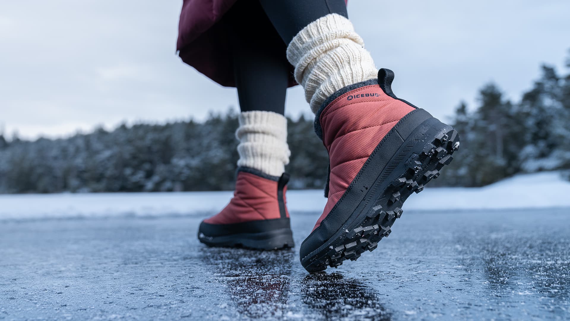 Close-up of a person walking on icy ground wearing red snow boots with thick soles and cream-colored leg warmers. Snowy landscape in the background.