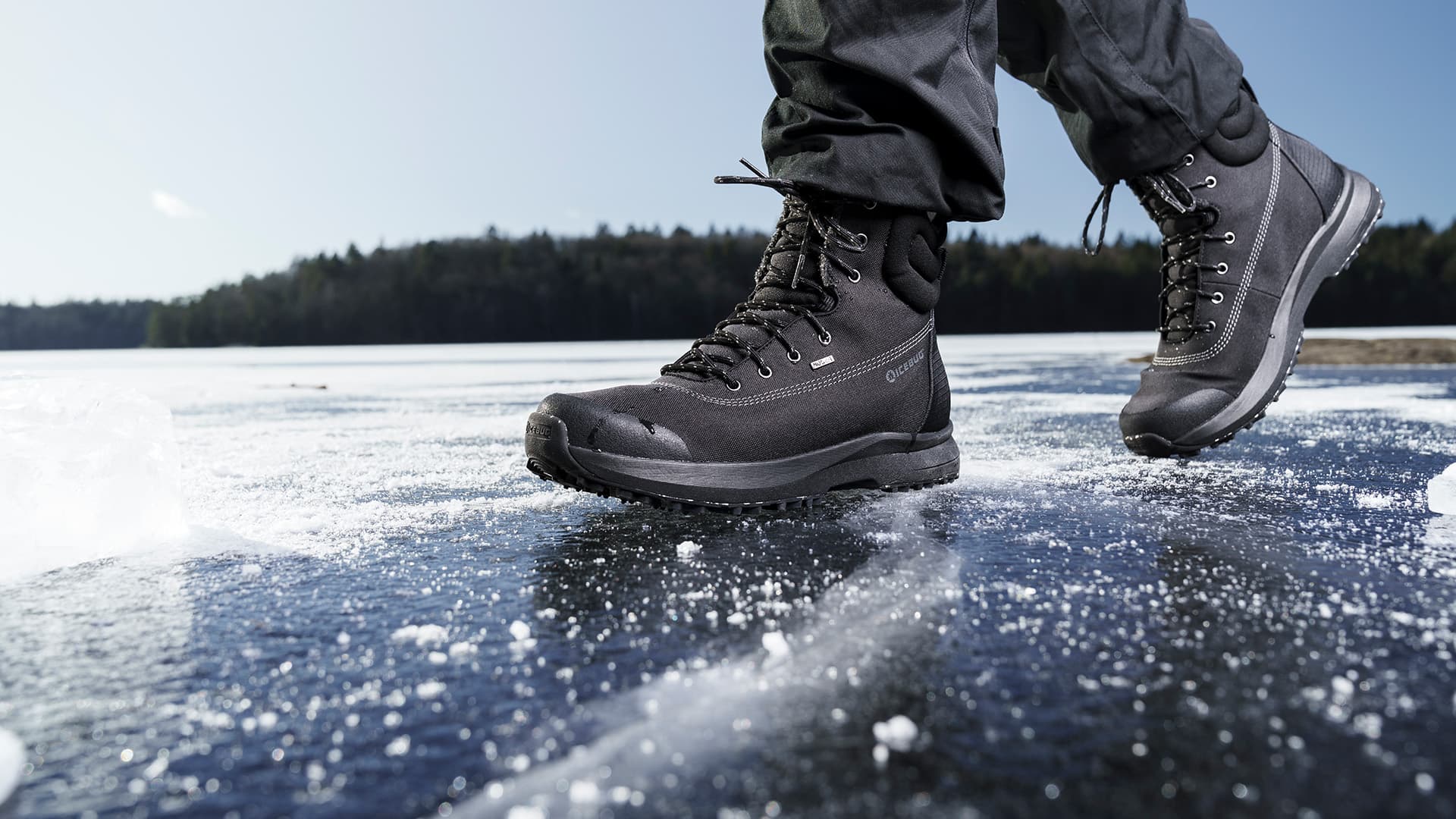 Close-up of a person wearing black hiking boots walking on a frozen, snow-dusted lake with a forest in the background.