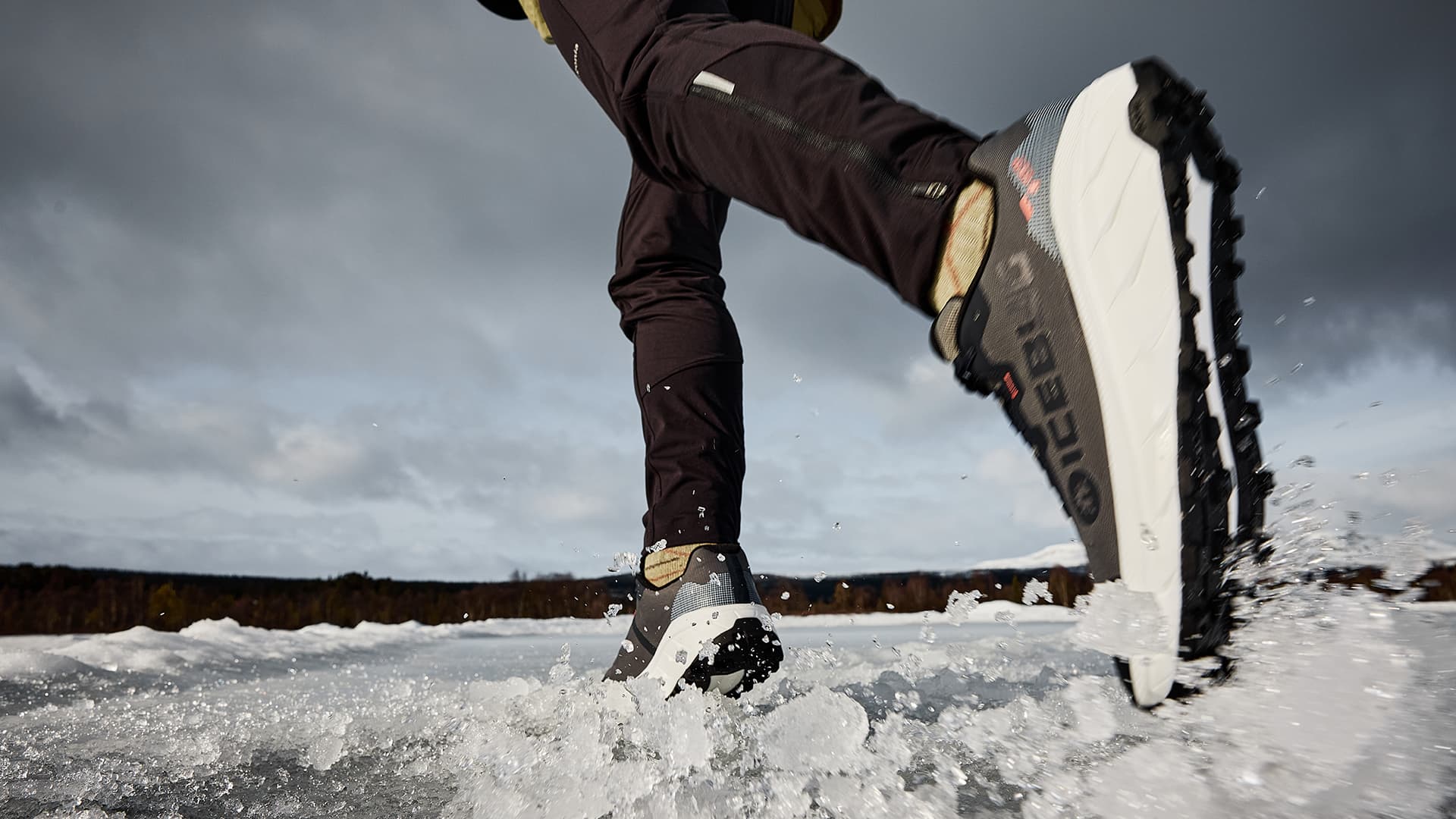 Close-up of a person running on ice wearing Icebug spiked shoes, kicking up snow. Overcast sky in the background.