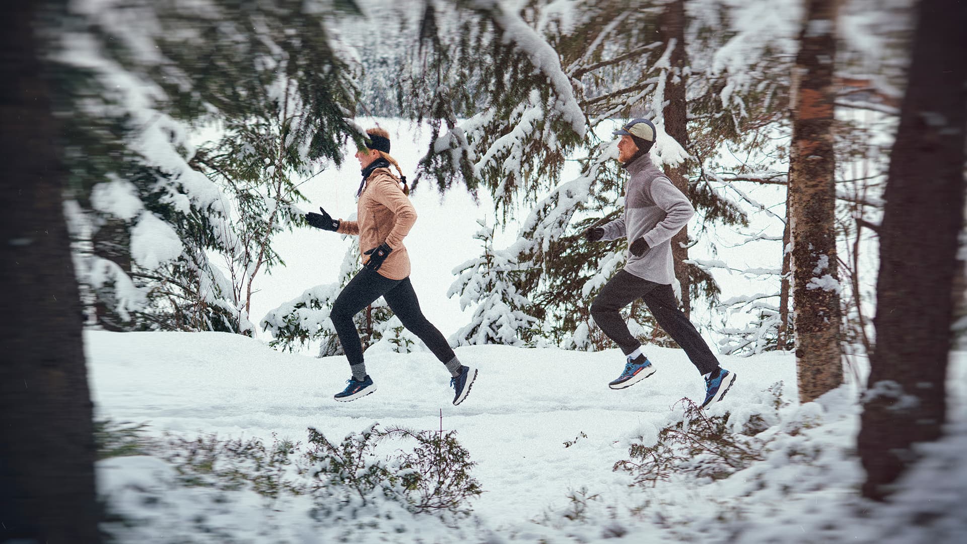 Two people jogging through a snowy forest, dressed in winter attire, surrounded by snow-covered trees.