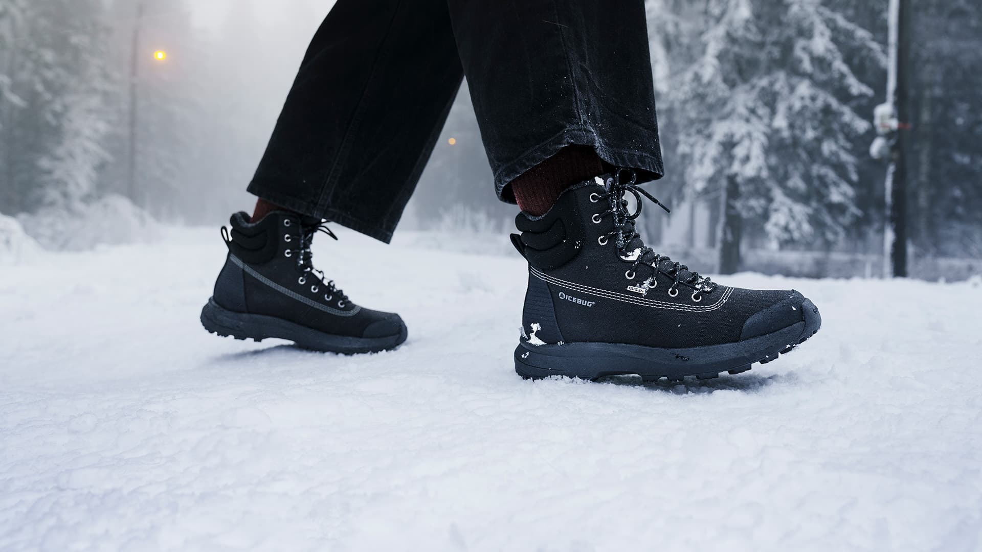 Person walking in snow wearing black boots and dark pants, with snow-covered trees and a streetlight in the background.
