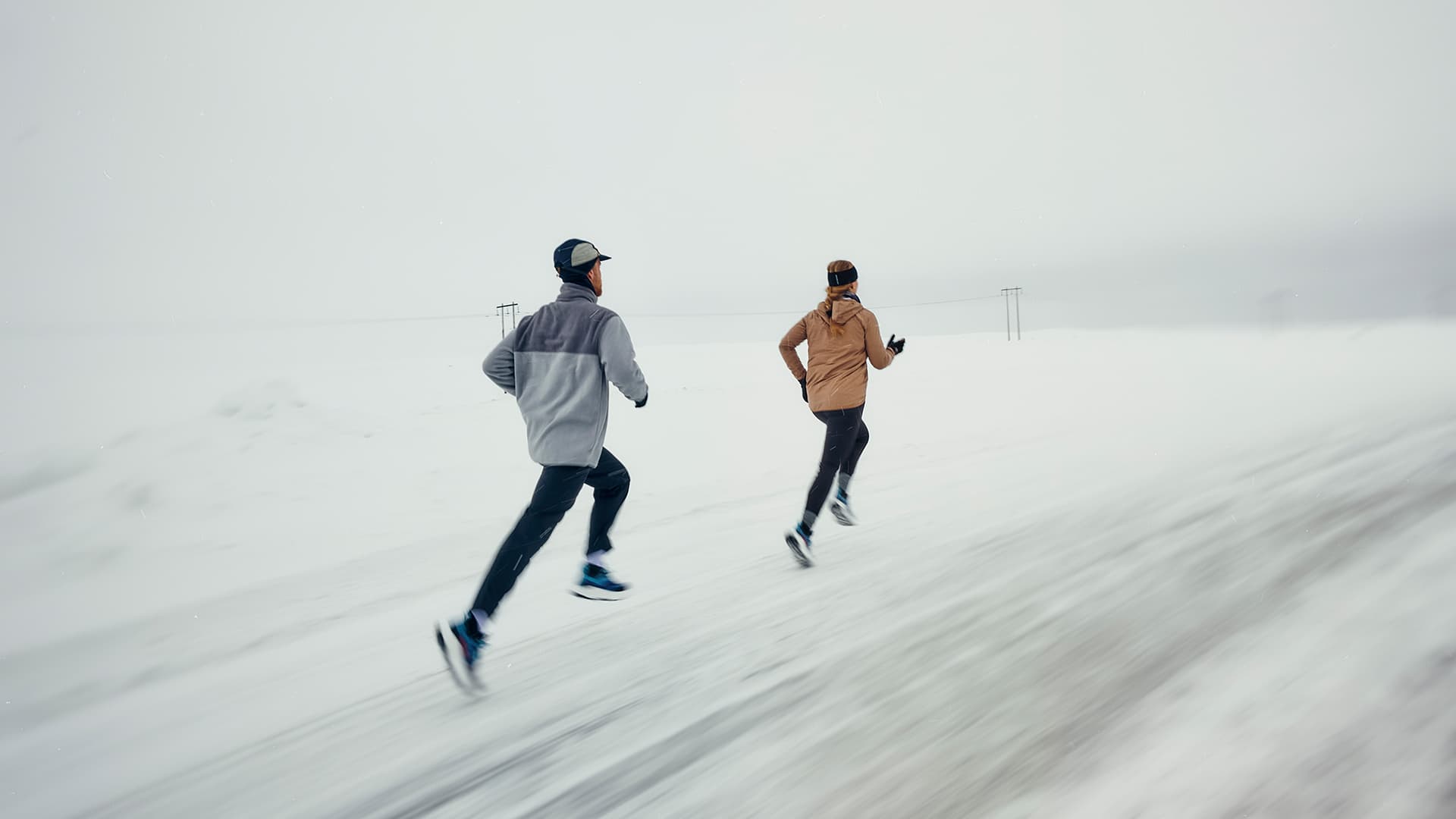 Two people running on a snowy road, wearing winter athletic gear. The landscape is overcast and covered in snow.