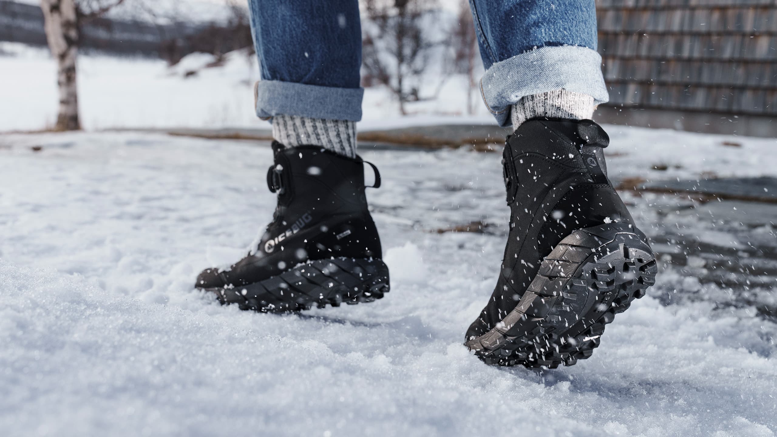 Person wearing black Icebug winter boots and rolled-up jeans walking on snow, with snowflakes falling around.
