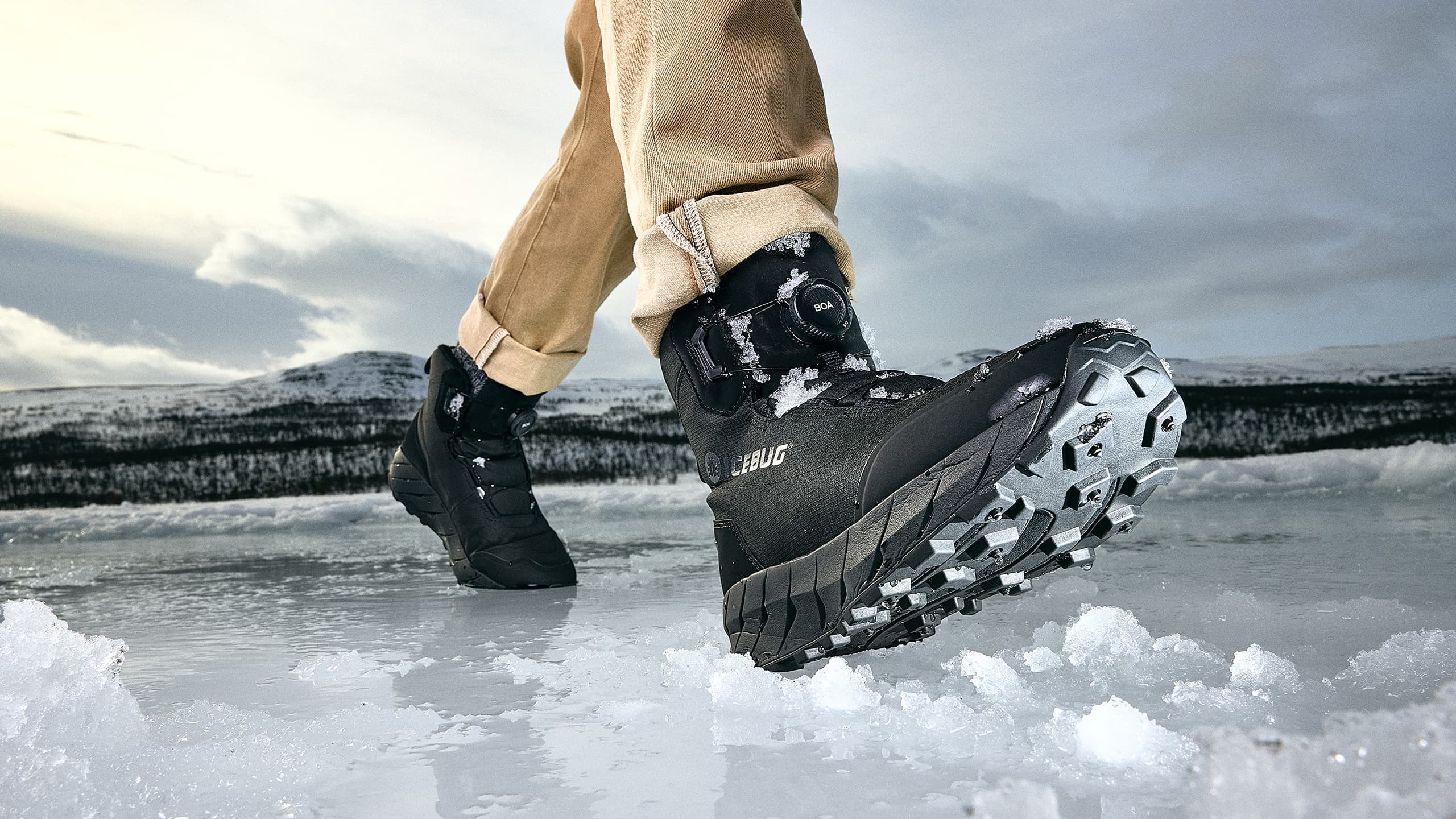 Close-up of a person wearing sturdy black winter boots from Icebug walking on an icy surface, with snowy mountains in the background under a cloudy sky.