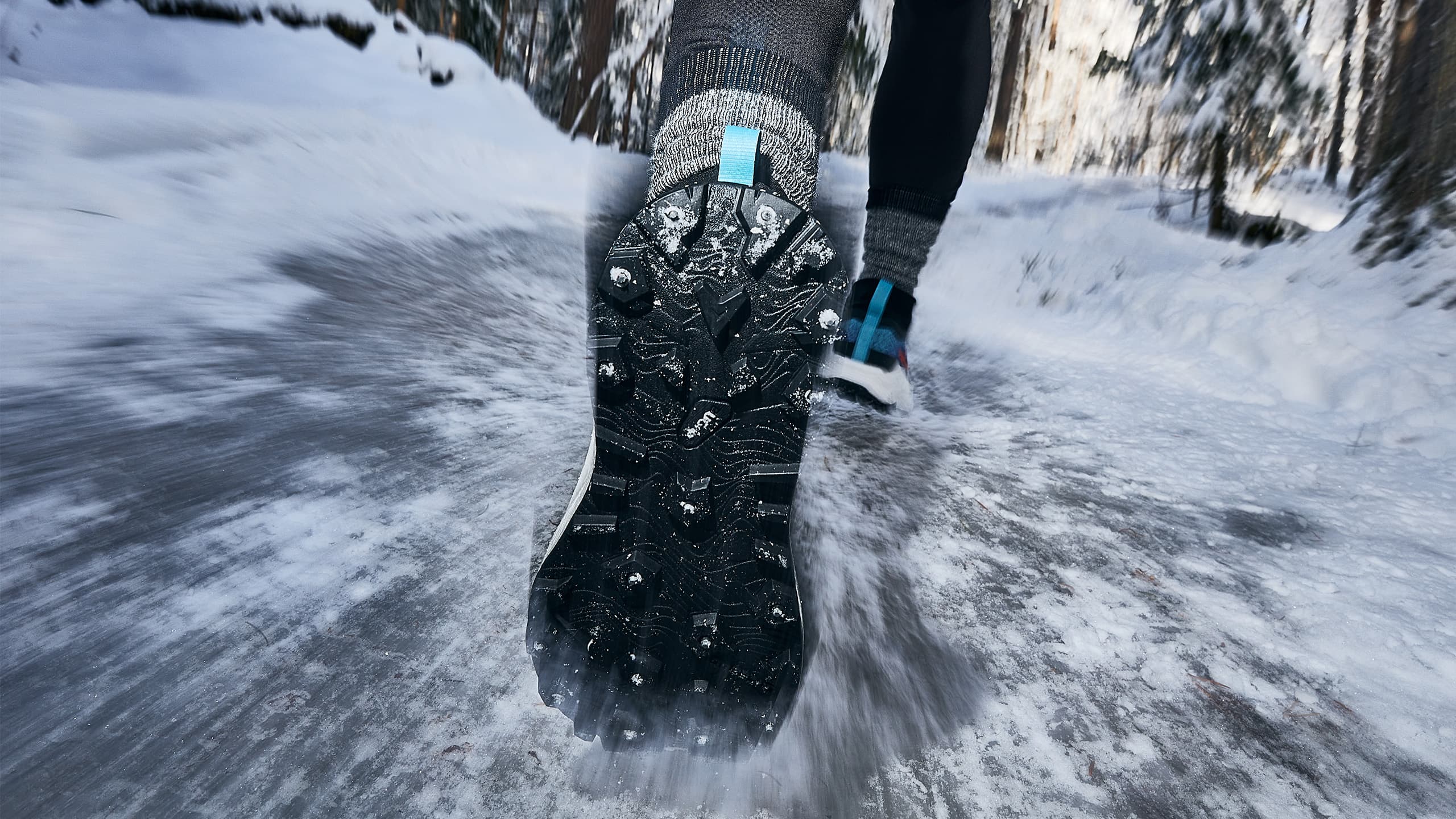 Close-up of a person running on a snowy trail, wearing studded winter running shoes for traction. Snow-covered trees line the path in the background.