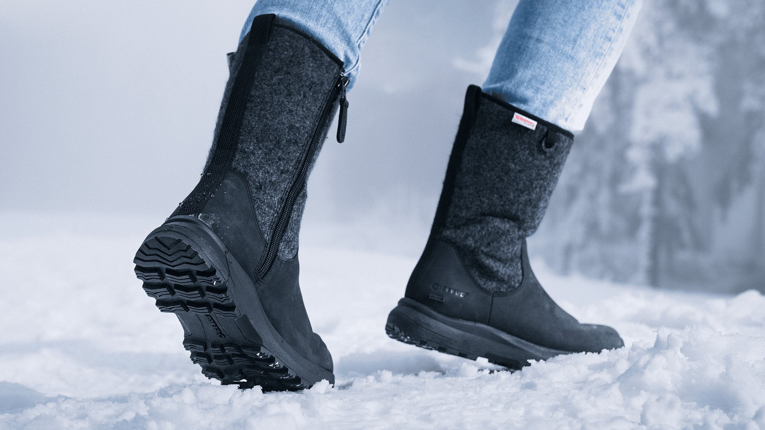 Close-up of a person wearing black winter boots and blue jeans, walking on snow. The boots have textured soles and a side zipper.