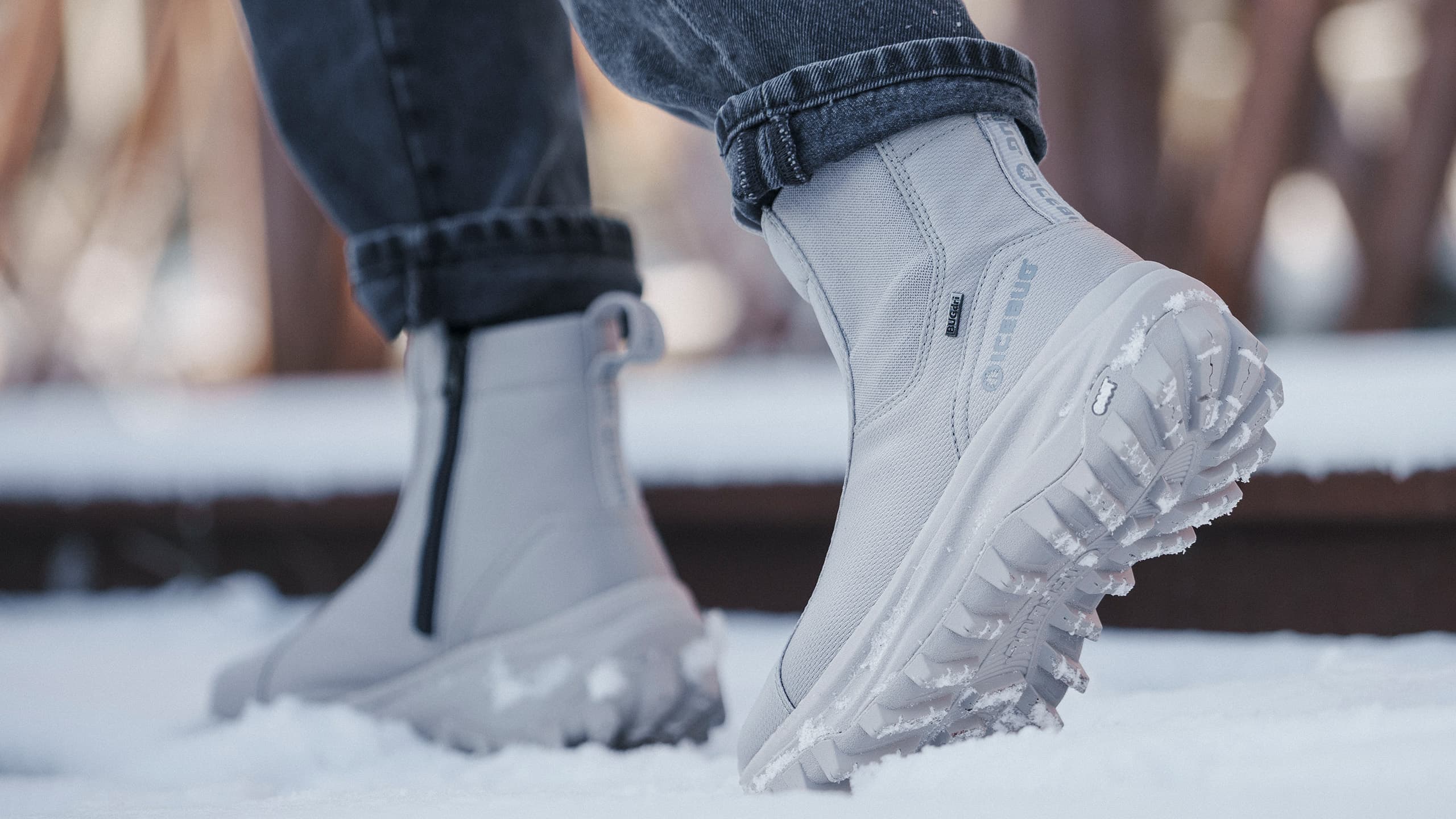 Close-up of a person wearing gray winter boots with rugged soles, walking on snow. The boots have zippers and the person is in jeans.