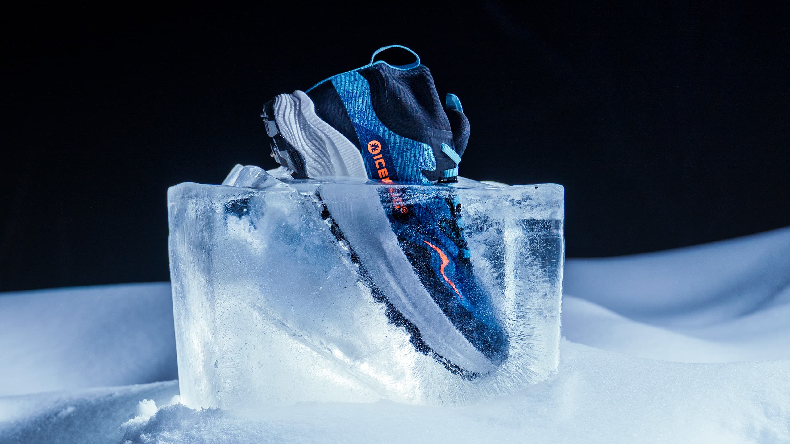 Blue and black athletic shoe encased in a clear ice block, surrounded by snow, against a dark background.