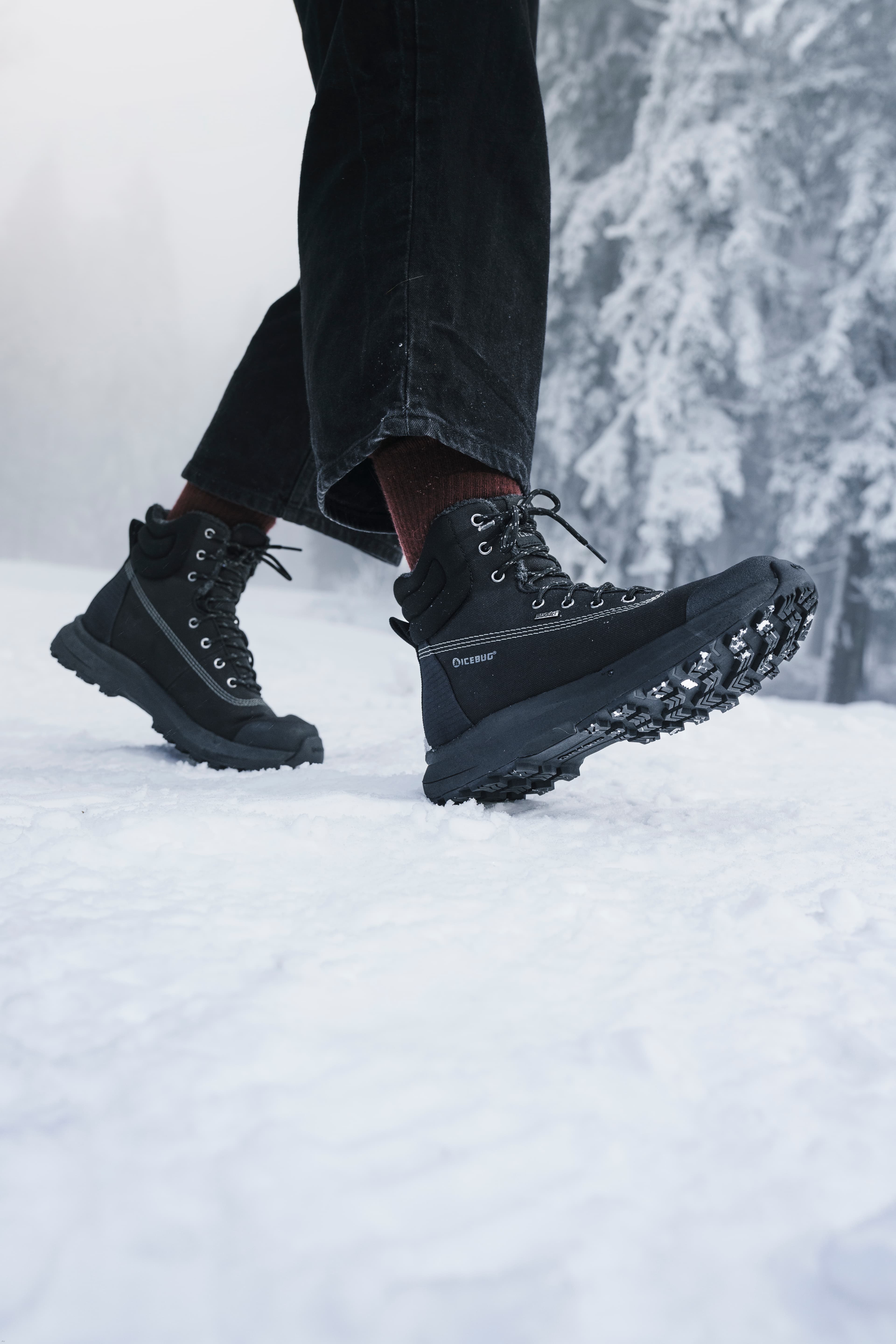 Person wearing black hiking boots walking on snow with snow-covered trees in the background.